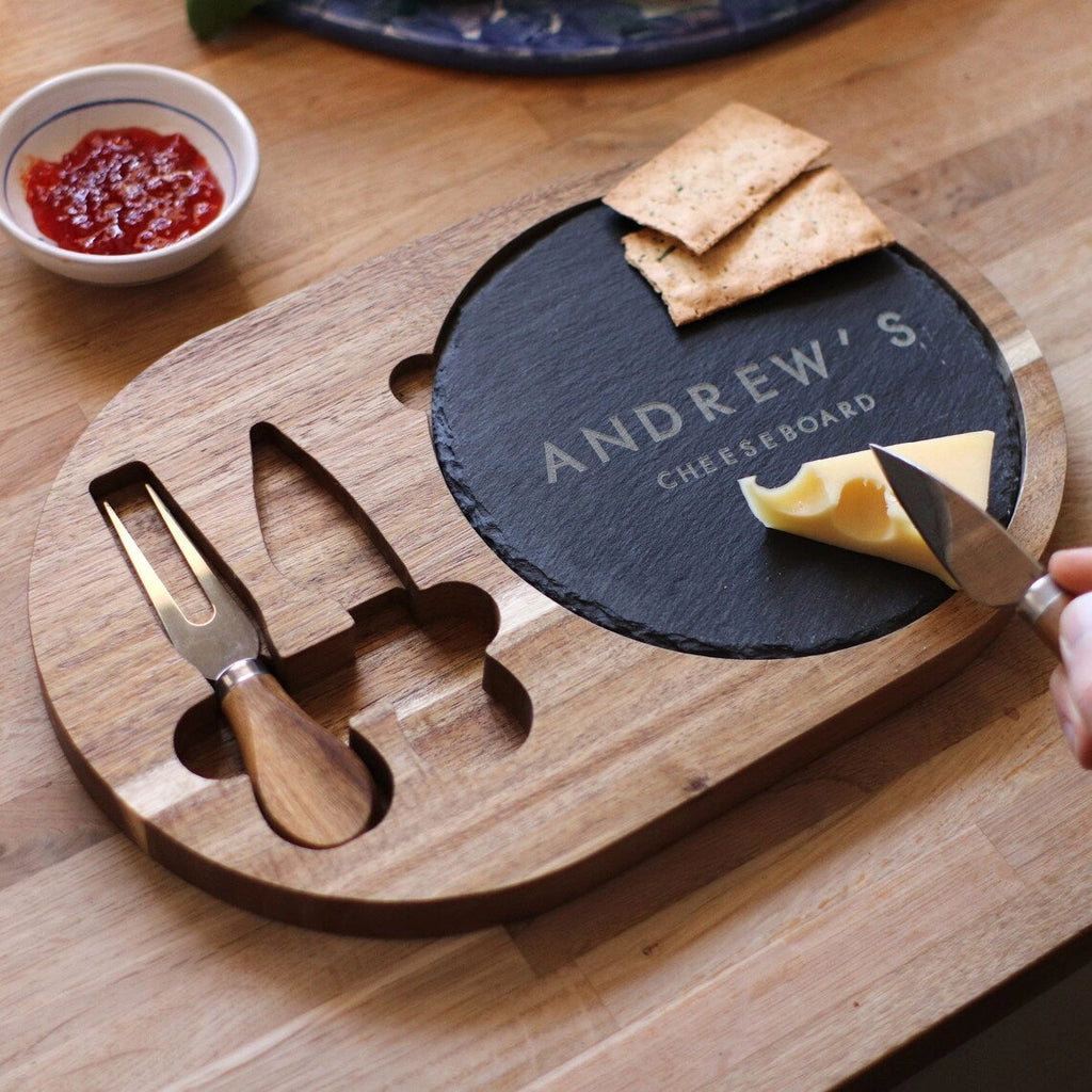 A wooden cheese board with carved slots holding a cheese knife and a small spreader, set on a wooden table. A round black slate insert reads 'ANDREW'S CHEESEBOARD' and is being used with crackers and cheese, with a hand holding a knife at the right edge.